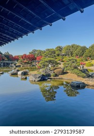 Viewed from beneath the dark lacquered eave of the main pavilion at Daisen Park, Sakai, Osaka, a large central garden island with layered sculpted pines, rounded topiary, and dark rocks is reflected in the still blue pond. Red maples and a small hillside gazebo are visible in the mid-distance, with the arched bridge and autumn trees completing the panorama beyond. A wide, airy composition with exceptional sky space and strong architectural framing.