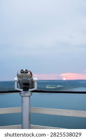 Viewing binoculars on an observation deck overlooking a lake at sunset.