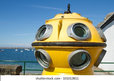 The Viewing Portals Of A Vintage Diving Bell Painted Yellow With Winch Attachments The Sea And Blue Sky In The Background.