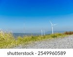 Viewpoint with gravel and grass growing on the green edge. Wind turbines tower over a lush forest. Background of mountain far away under blue sky.  Landmark of Khao Yai Thieng.