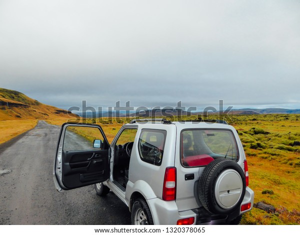 Vik/Iceland- 10 27 2018: A three door car pulled on\
the side of the road. The off-road car, 4W car. The driver\'s door\
are open. The road is graveled, leading through lava fields.\
Mountains around.\
\

