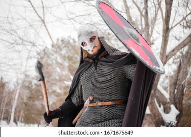 Viking In Winter With A Spear And A Round Shield Of Red-black Color. A Guy In A Helmet And Chain Mail In The Snow. A Man In Armor Goes To Battle