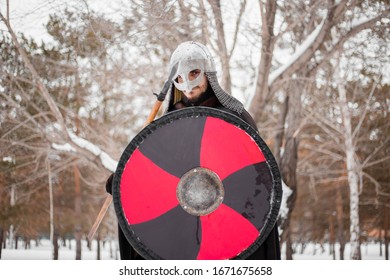 Viking In Winter With A Spear And A Round Shield Of Red-black Color. A Guy In A Helmet And Chain Mail In The Snow. A Man In Armor Goes To Battle