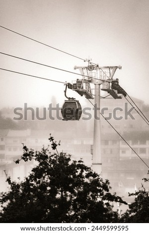 A Vilanova de Gaia cable car gondola suspended on hanging steel cables ascending under a cloudy sky with the classic Gaia neighborhood shrouded in fog. Black and white.