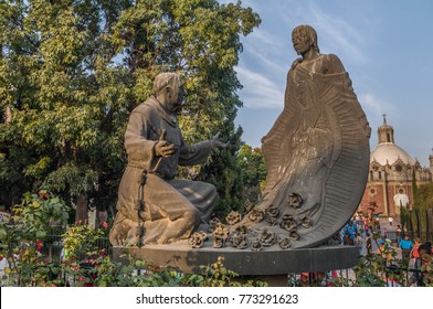 VILLA OF GUADALUPE, MEXICO CITY, DECEMBER 02, 2017. Sculpture Of Juan Diego And Fray Juan De Zumarraga In The Gardens Of The Villa Of Guadalupe. At The Background We Can See The Pocito Church. 