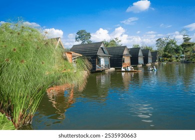 villa on the lakeside with two people rowing a small boat in the water. Clear sky, green trees, and calm water reflection - Powered by Shutterstock