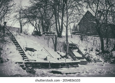 Village House On High River Bank. Ice And Snow Covered Water Surface. Slippery Stairs To Reach Water, Boat On Shore.