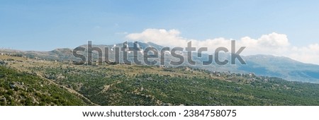 A village sits on a hill ridge across a rocky landscape, with Mount Lebanon mountain range, snow-covered in spring, a popular hiking destination in Lebanon, Middle East