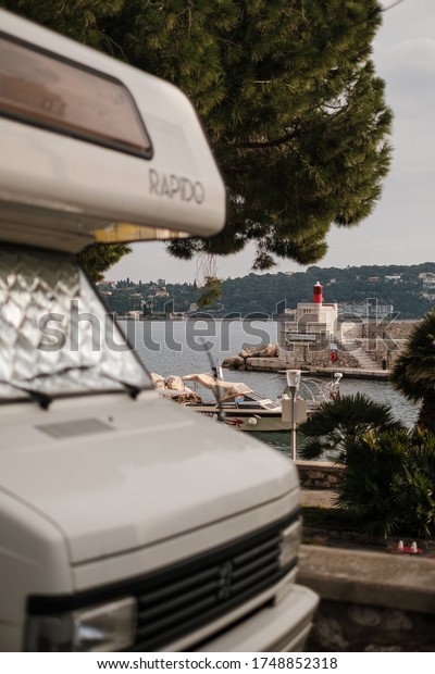 In Villefranche sur\
Mer a camping car is parked in the street with the port and the\
lighthouse of the city in the background. Villefranche sur Mer,\
France, January 07, 2020.