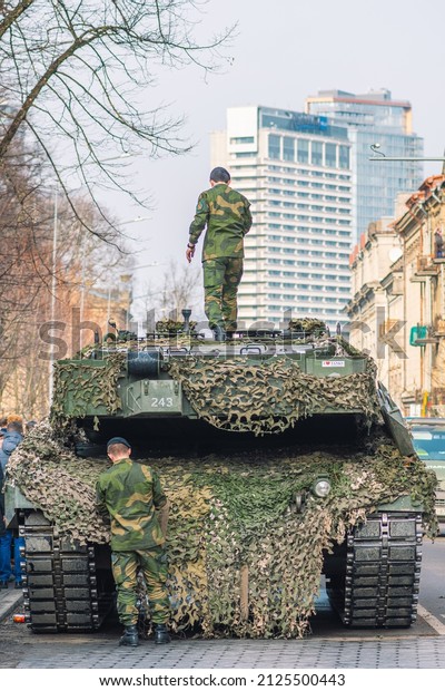 Vilnius, Lithuania - February 16 2022: NATO\
response force. Armored tank with cannon and camouflage coating\
with soldiers and other vehicles on the road of a city with\
skyscrapers on\
background