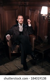 Vintage 1900 Fashion Man With Beard. Sitting In Old Wooden Reading Room. Smoking A Cigar.