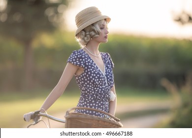 Vintage 1930s fashion woman in blue summer dress standing with bicycle in rural landscape.