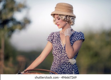 Vintage 1930s fashion woman in blue summer dress standing with bicycle in rural landscape.