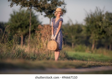 Vintage 1930s Summer Fashion Woman With Blue Dress And Straw Hat Standing With Handbag On Rural Road.
