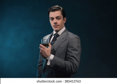 Vintage 1940s Fashion Man In Suit And Tie Holding Glass Of Whiskey Against Blue Wall.