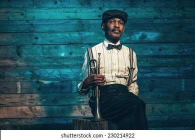 Vintage african american senior jazz musician with trumpet in front of old wooden wall. Wearing suit and cap.