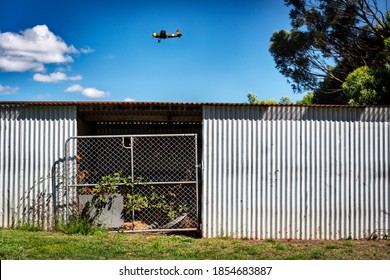 A vintage aircraft flying over a silver corrugated shed with a tin roof and metal gate overgrown with vine against a blue sky with clouds - Powered by Shutterstock