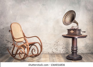Vintage antique gramophone phonograph turntable with brass horn on wooden table and aged rocking chair front concrete wall background with shadow. Retro old style filtered photo 