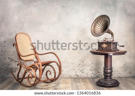 Vintage antique gramophone phonograph turntable with brass horn on wooden table and aged rocking chair front concrete wall background with shadow. Retro old style filtered photo 