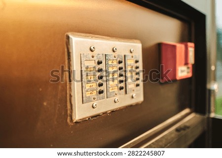 A vintage apartment building intercom panel with numbered brass labels and black buzzer buttons. A red fire alarm pull station sits beside it. Warm light catches the worn metal surface.
