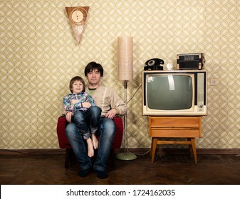 Vintage Art Portrait Of Litle Boy With His Father Sitting On Armchair In Room With Interior From 70s 20th Century, Retro Stylization, Image Toned. Family At Home With Old Tv Set