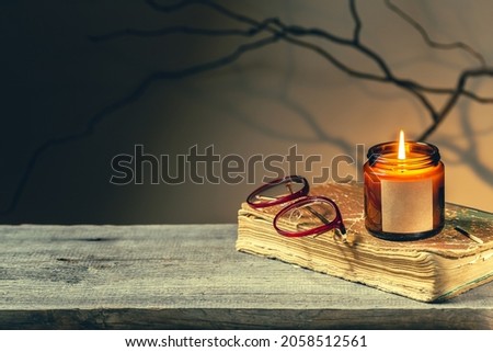 Vintage background with an old antique book, glasses and candle in a glass jar burning at night. Tree brunches and shadows make spooky dark academia atmosphere. Cozy fall season reading time