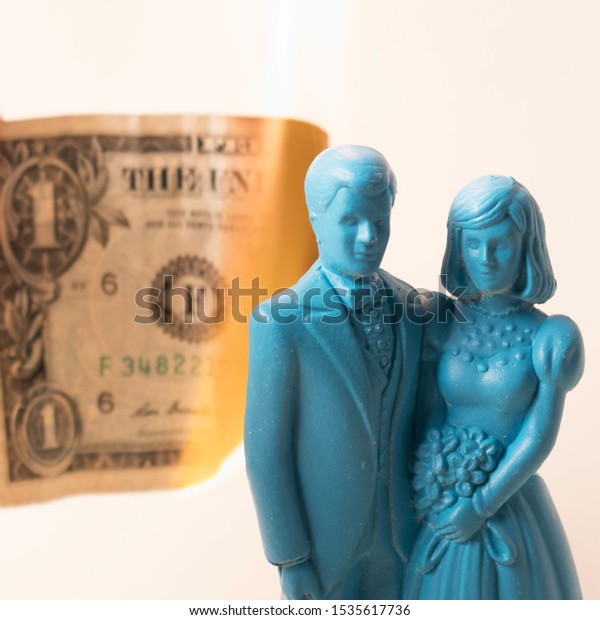 Vintage blue wedding cake topper couple in front\
of a dollar bill on fire. Weddings can be costly and a waste of\
money. Couple fighting over finances. Man and wife in financial\
trouble or divorce.