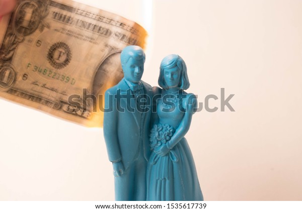 Vintage blue wedding cake topper couple in front of a\
dollar bill on fire. Weddings can be costly and a waste of money.\
Couple fighting over finances. Man and wife in financial trouble or\
divorce. 