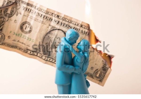 Vintage blue wedding cake topper couple in front
of a dollar bill on fire. Weddings can be costly and a waste of
money. Couple fighting over finances. Man and wife in financial
trouble or divorce.