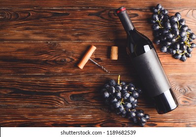 Vintage bottle of red wine with blank matte black label, corkscrew & bunches grapes on wooden table background. Expensive bottle of cabernet sauvignon concept. Copy space, top view, flat lay.