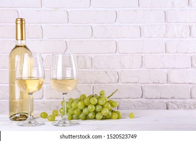 Vintage bottle of white wine without label, two glasses and bunch of grapes on wooden table, lofty white brick wall background. Expensive bottle of chardonnay concept. concept. Copy space.