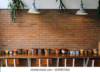 Vintage Brick Wall Background, Ceiling Lamps, Green Plants, Colorful Empty Clay Flower Pots On Wooden Table And Chairs In A Coffee Shop.