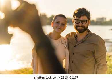 Vintage camera. Black male photographers POV. Blurred foreground. Caucasian couple - bald woman and bearded man - in casual clothes smiling and posing during photoshoot. Golden hour. River in the - Powered by Shutterstock