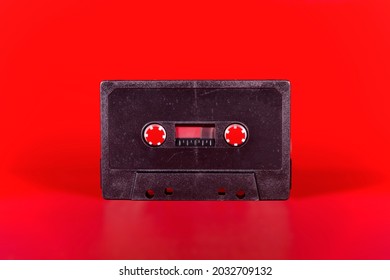 A vintage cassette tape from the 1980s era (obsolete music technology), dark gray plastic body without any label or text, isolated on a shiny red backdrop.
