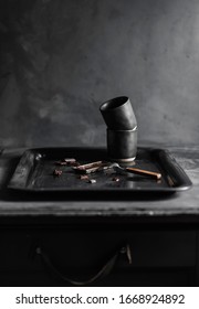 A vintage chocolatebar mold filled with melted dark chocolate and some chocolate chunks and a stack of black coffeecups on a vintage tray against a dark grey background