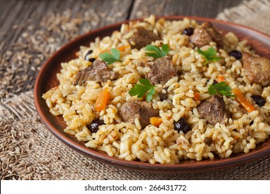 Vintage clay bowl with traditional spicy food called pilaf, on wooden background. Cooked with fried lamb, rice, garlic, carrot and raisins.