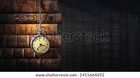 Vintage clock hanging on a chain on the background of old books. Old watch as a symbol of passing time. Concept on the theme of history, nostalgia, old age. Retro style.