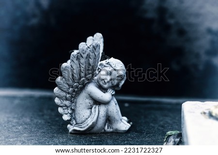 Vintage dirty statue of weep angel on tomb stone in old cemetery. Small ancient winged angelic figurine on dark sadness grave yard. Baby cherub figure symbol of consolation, grieve, belief protect