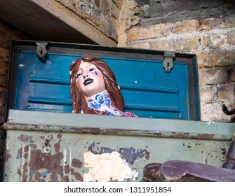 Vintage Doll Head With Long Brown Hair, Black Lips, Blue Rose Tattoo And A Blue Tear Drop Under The Left Eye Sits Inside An Old Metal Trunk With Blue Rusty Lid.