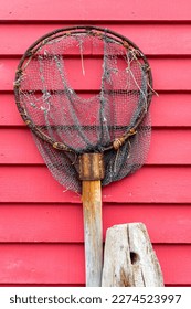 A vintage fishing dip net hanging on a vibrant red wooden horizontal clapboard wall. The scoop is rusty and the cloth mesh rope net is torn and worn with holes. The equipment is grungy.