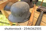 Vintage German Stahlhelm helmet from World War I or II on a wooden surface, accompanied by a spiked trench club and a field shovel with leather straps, evoking a wartime setting.