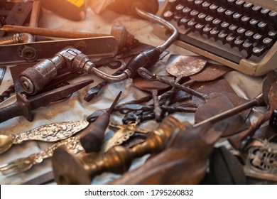 Vintage Hand Crank Brace Drill Wood Handle,  Rusty Metal Parts, Silver Spoon And Fork, Worn Typewriter On Countertop At Antique Bazaar.