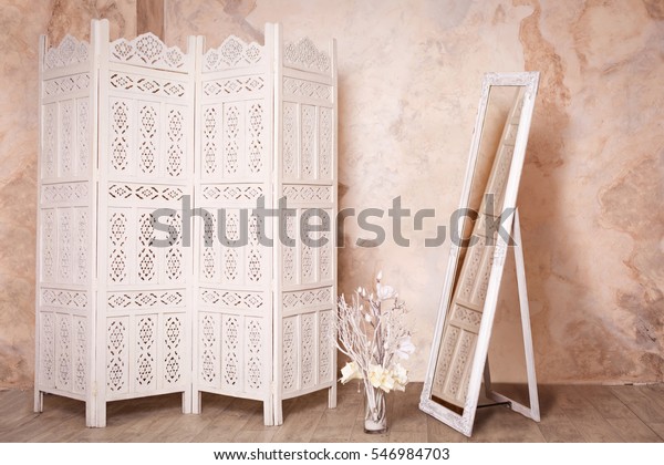 Vintage\
interior with white delicate decorative wood panel and mirror on\
brown plaster wall. Boudoir wedding room. Retro folding screen and\
mirror. Vintage ornate carved folding screen.\
