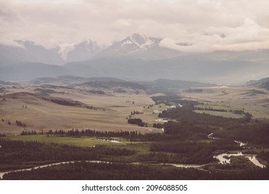 Vintage landscape with vast plateau with mountain river and forest on background of snowy mountain ridge under cloudy sky. Mountain valley and mountain range among low clouds in sepia and faded tones.