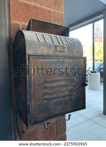 Vintage looking mailbox. Rusty, old, and black looking mailbox against a brick wall. A metal mailbox that say “Letters” on the top. Outside with open space in the background.