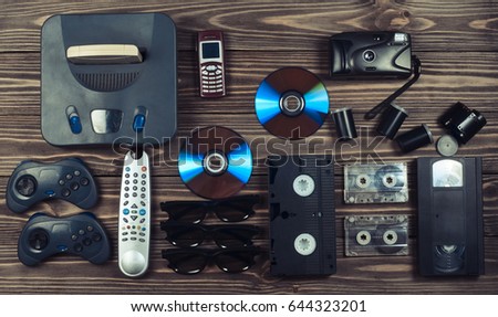 Vintage media and information technology. Entertainment 90s. Game console, gamepads, disks, audio cassettes, video cassettes, phone, film camera on a  wooden table. Top view. Flat lay. 