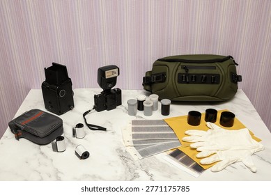 a vintage medium format camera a flash film rolls negatives in sleevers white gloves and a photography bag are laid out on a table preparing the workflow in an analog darkroom for the photographer