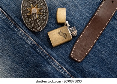 Vintage metal lighter and leather belt with massive buckle on denim with double stitching. Illustrative editorial image. Ukraine, Zhytomyr, December, 13, 2021