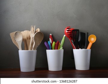 Vintage and modern kitchen hand tool utensils such as spoon, fork, spatula, ladle and knife , made from wood and plastic, in 3 white pots on a wood counter in front of dark grey concrete plaster wall.