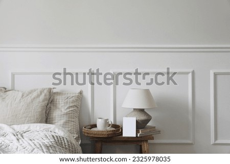Vintage neutral classic bedroom interior. Cup of coffee on wicker tray, table lamp on wooden bench. Blank greeting card, invitation mockup. White wall background. Gingham bedding set, pillows.